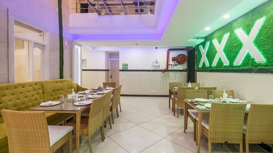 A restaurant interior with a long tufted sofa, wooden tables and chairs, a decorative wall design, and a tiled floor, lit by soft lighting. - Hotel Diamond Plaza, Chandigarh.