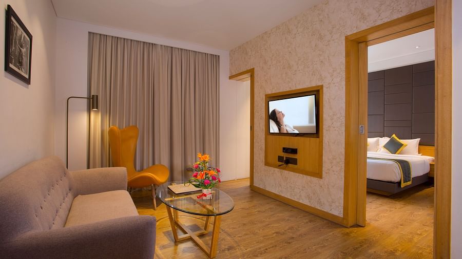 view of a living room with sofas and a small table, all facing a television mounted on the wall and a door leading to the bedroom on the right - Hotel Fountain Tree By TGI - JP Nagar, Bangalore