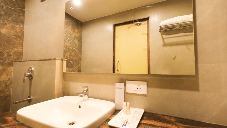 Bathroom counter featuring a white ceramic washbasin, large mirror, wall light, and complimentary toiletries tray at Hotel Grand Sabarees Madurai.