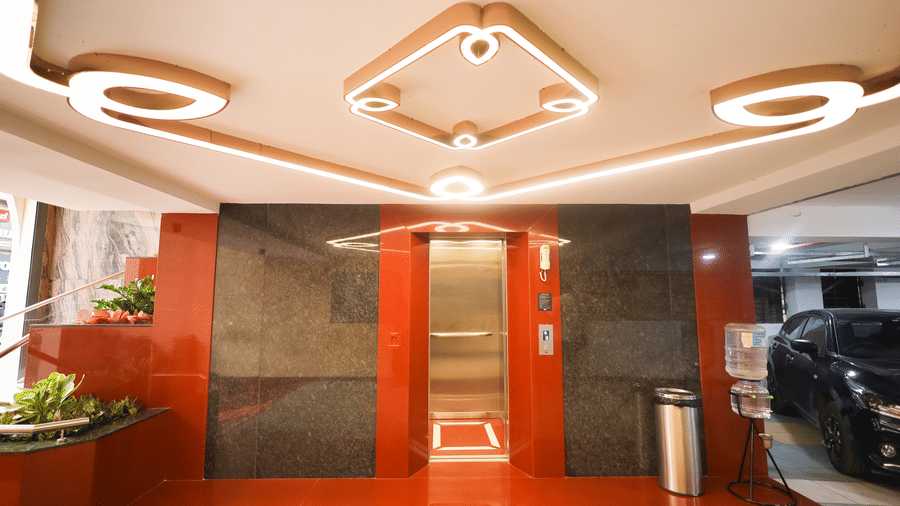 Glass elevator entrance with red panel frame, ceiling lights, and indoor plants in the lobby at Hotel Grand Sabarees Madurai.