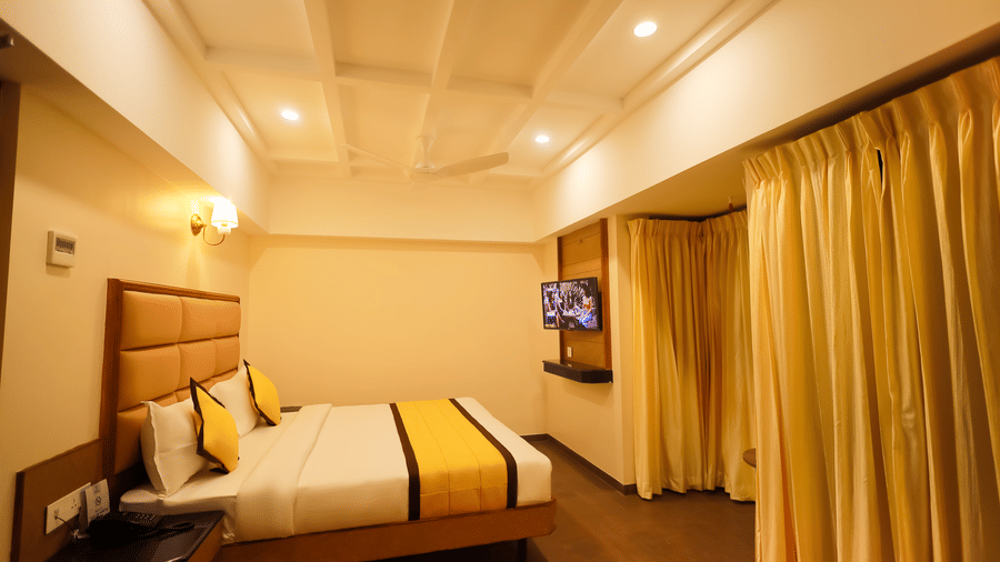 Bedroom view at Hotel Grand Sabarees Madurai showing a double bed, bedside lights, yellow window curtains, and a wall-mounted TV.