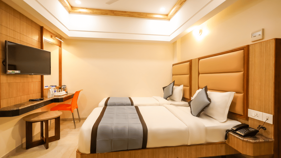 Twin guest room at Hotel Grand Sabarees Madurai showing two single beds, wall-mounted television, work counter, chair, and telephone.