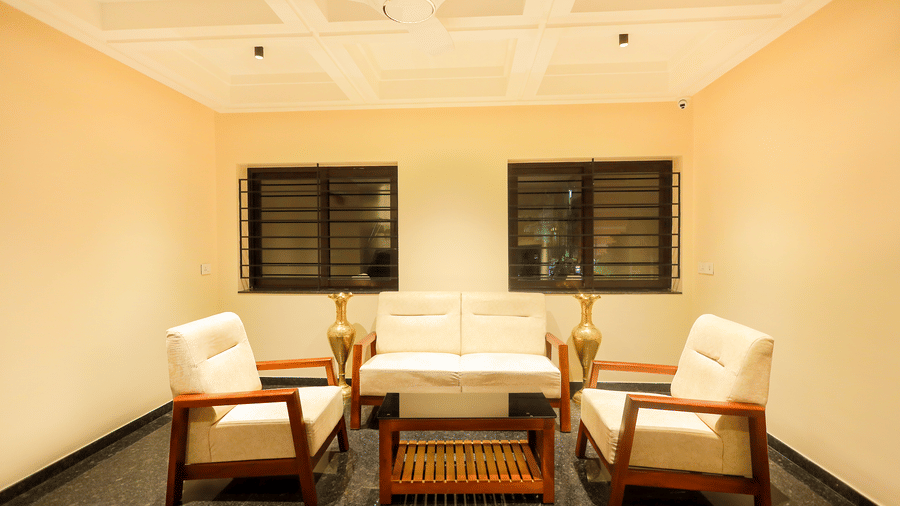 Seating area at Hotel Grand Sabarees Madurai with a light-colored sofa, two armchairs, a wooden coffee table, and two windows.