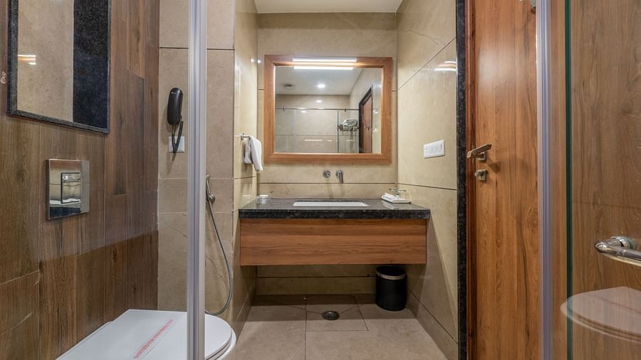  A well-lit bathroom at Hotel Highway King, Bagru, showing a vanity with a dark countertop, a sink, a large mirror, and a wooden door.