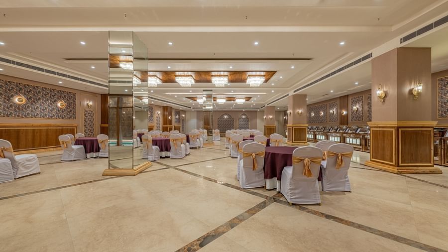 Spacious banquet hall at Hotel Highway King, Bagru, with arranged chairs, decorative ceiling panels, and wood-paneled walls.