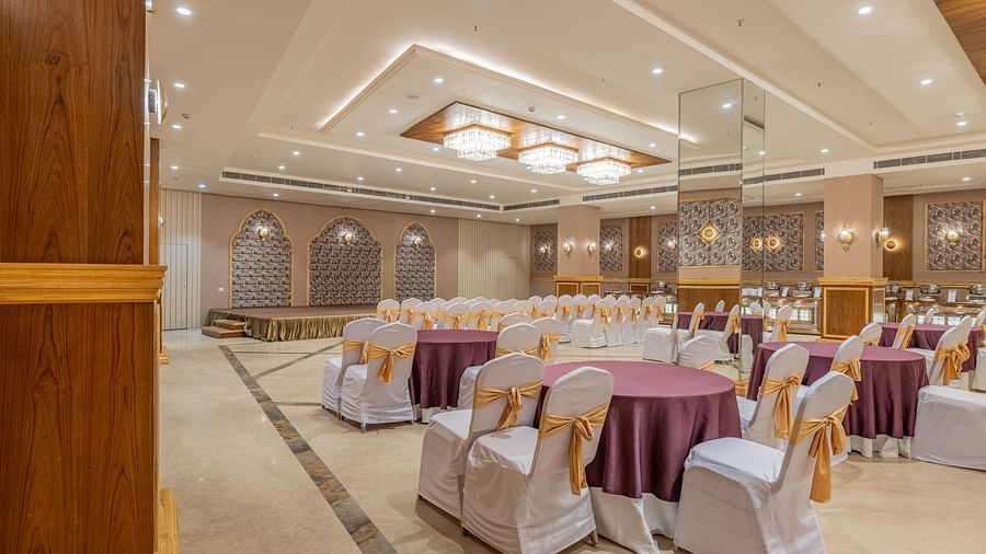 Banquet hall at Hotel Highway King, Bagru, with round tables, decorated chairs, chandeliers, and decorative wall panels.