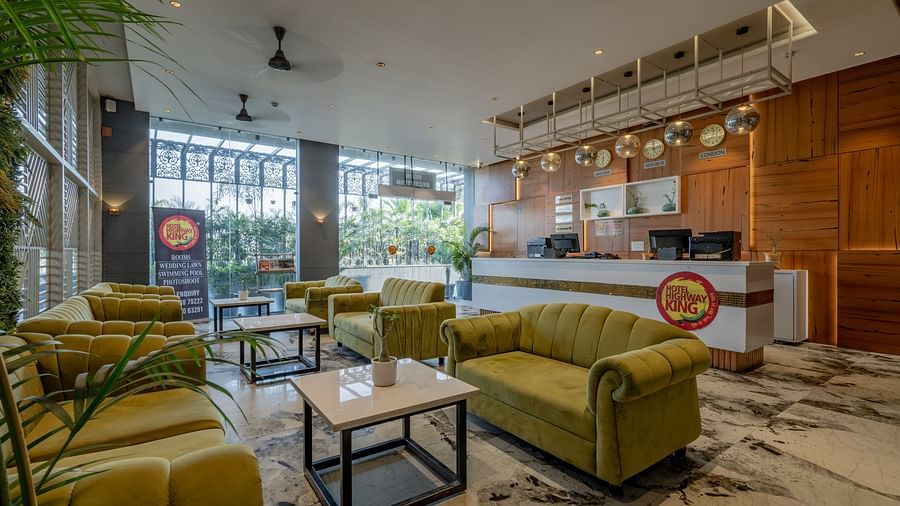 Lounge and reception area at Hotel Highway King, Bagru, featuring sofas, tables, large windows, and wooden paneled walls.