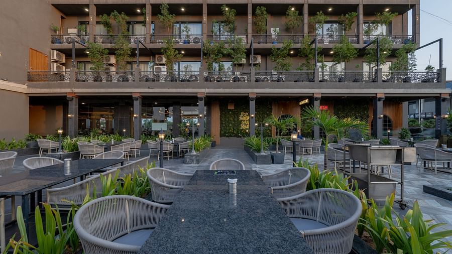 Outdoor dining area at Hotel Highway King, Bagru, with tables, chairs, plants, and multi-storey hotel building in the background.