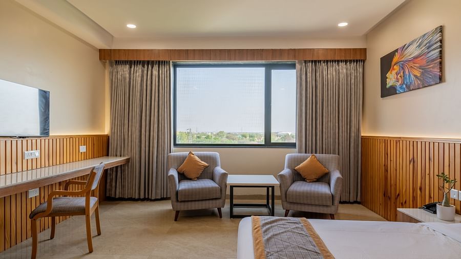 A view of a club room at Hotel Highway King, Bagru, with a double bed, a large window offering natural light, a seating area with two armchairs, and a colorful artwork.