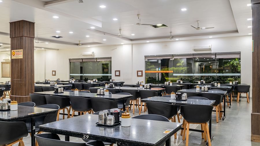 Extended view of the restaurant at Hotel Highway King, Behror, furnished with dining tables with varying seating capacities, ample lighting and windows that offer views of the trees outside.