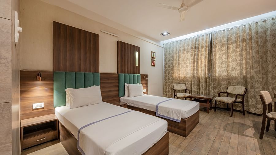 Twin beds with upholstered headboards and bedside tables beside a seating area for two and drawn curtains in the Maharaja Deluxe Rooms of Hotel Highway King, Behror.