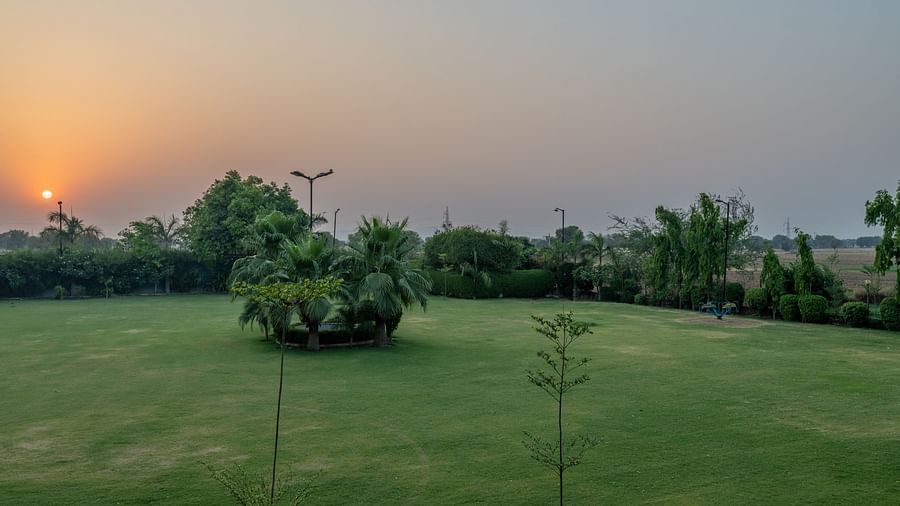 An extended view of the expansive open garden area in the premises of Hotel Highway King, Behror, seen with trees, grass, and glowing sun in the background.