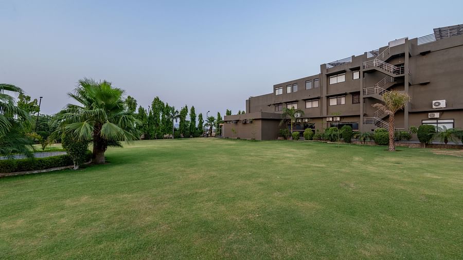 An extended view of the expansive open garden area in the premises of Hotel Highway King, Behror, seen with trees, grass, and the hotel's structure in the background.
