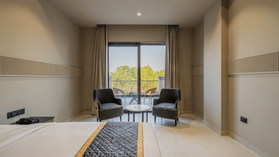 Hotel bedroom looking out towards a glass balcony door with 2 armchairs, a round table, and distant green trees at Hotel Highway King, Harmada.