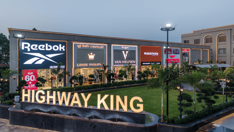 A decorated green lawn, a stone wall with illuminated lettering, and large retail brand signboards on the building facade at Hotel Highway King, Harmada.