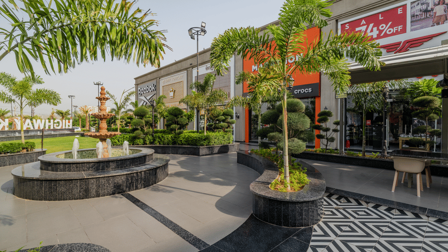 A stone fountain, retail shop entrances, and small palm trees at Hotel Highway King, Harmada.