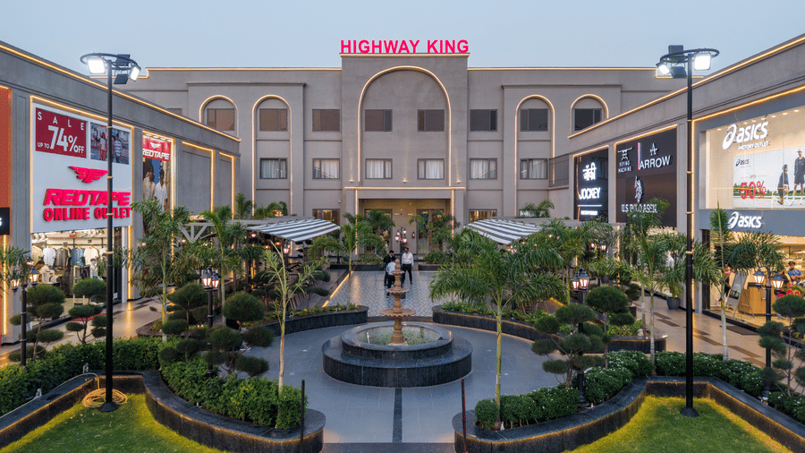 A central circular water fountain, manicured green lawns with palm trees, and retail storefronts under lights at Hotel Highway King, Harmada.