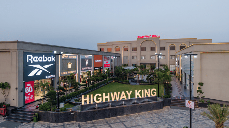 An illuminated name signage on a stone boundary wall, a neat garden lawn, and a retail building wing with brand logos at Hotel Highway King, Harmada.