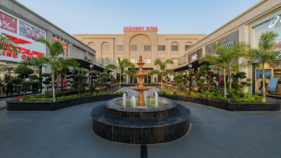A wide view showing a tiered fountain, a main building, and retail outlets at Hotel Highway King, Harmada.