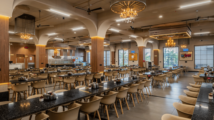 A dining hall showing rows of black tables, wooden pillars, and ceiling light fixtures at Hotel Highway King, Harmada.