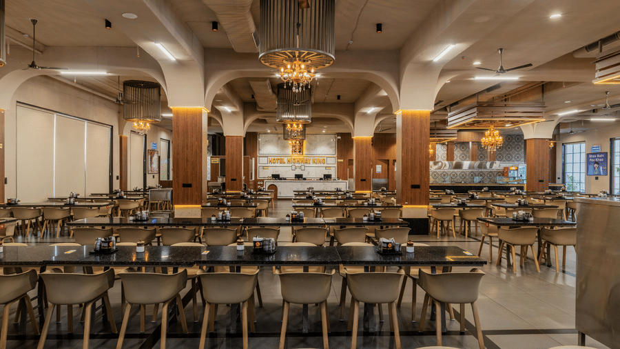 Long dining tables arranged in neat rows with chairs, pillars, and multiple ceiling fans visible at Hotel Highway King, Harmada.