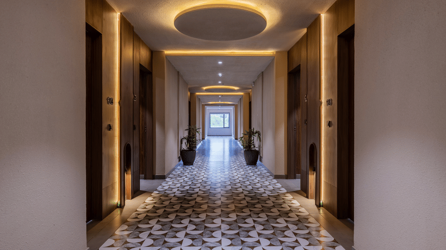 A long corridor with a large circular ceiling light, wooden room doors on both sides, and a patterned tile floor walkway at Hotel Highway King, Harmada.