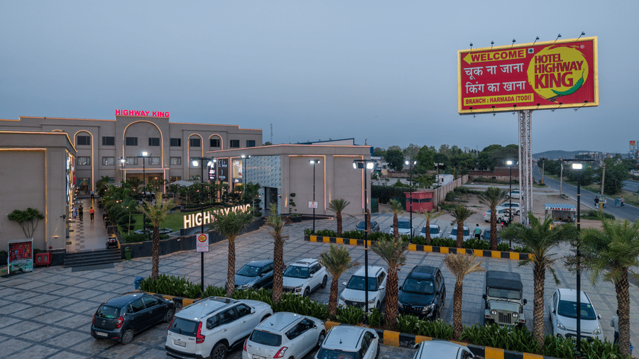 An open parking lot with multiple cars, a row of palm trees, and a tall advertisement board next to a road at Hotel Highway King, Harmada.