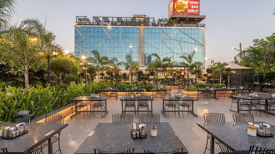 Outdoor seating area with multiple tables and chairs arranged in rows with greenery and the facade of Hotel Highway King at the background.