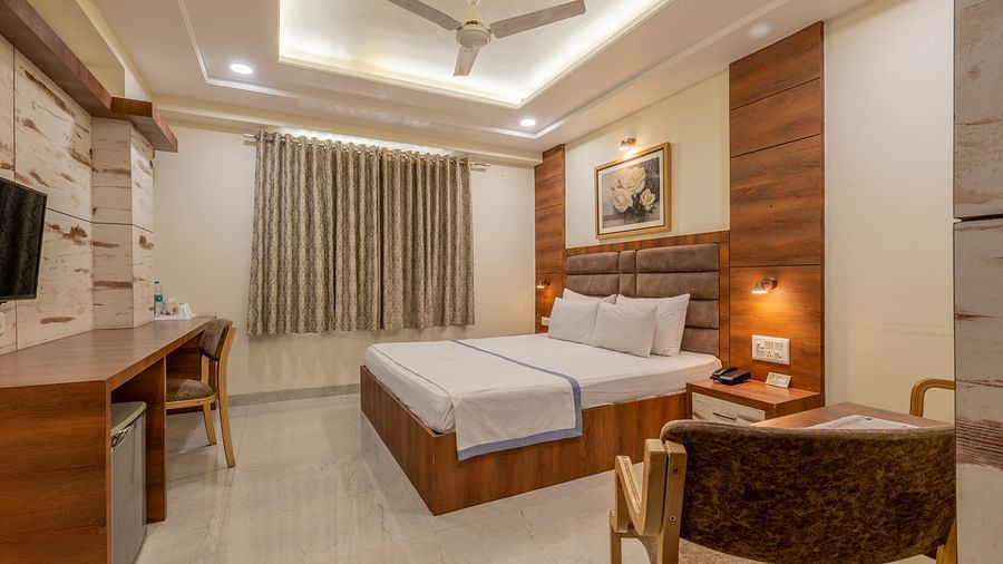 A room featuring a bed with white and blue linen, a wooden desk and chair, and a wall art at Hotel Highway King, Jaipur.