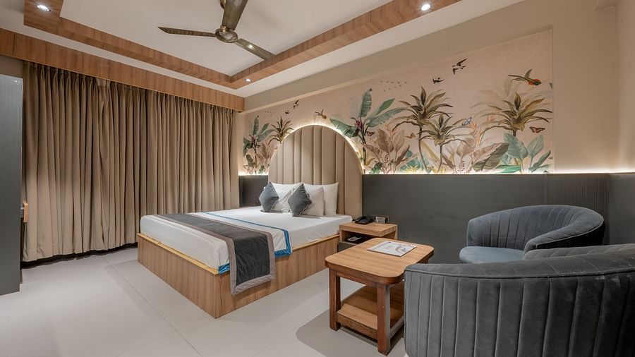 A wide view of the room with a king size bed, brown curtains, grey chairs, a wooden table and a flowery mural on the wall at Hotel Highway King, Jaipur.