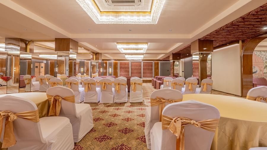 Round banquet tables covered with white cloth and gold sashes, set under a large chandelier in a decorated hall at Hotel Highway King, Jaipur.
