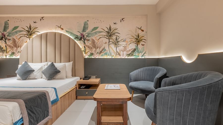 A partial view of a king size bed featuring grey accents, grey armchairs, a wooden table and plant-themed wall mural in a well-lit room at Hotel Highway King, Jaipur.
