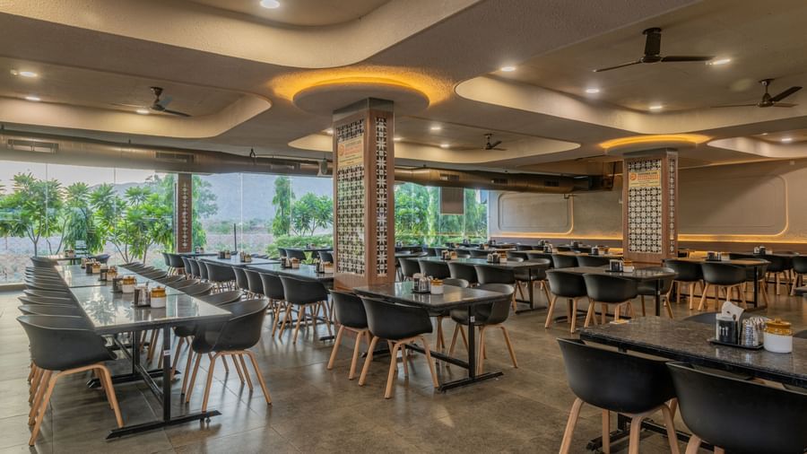 Long view of the restaurant interior at Hotel Highway King, Neelka, with neatly arranged tables and chairs, glass panels, and a lawn view in the background.