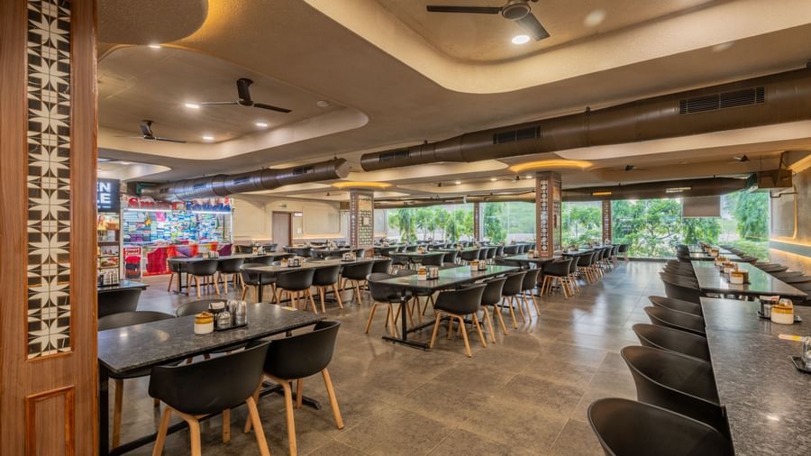 A diagonal view of the restaurant seating area, featuring panoramic glass panels in the background and warm lighting, at Hotel Highway King, Neelka.