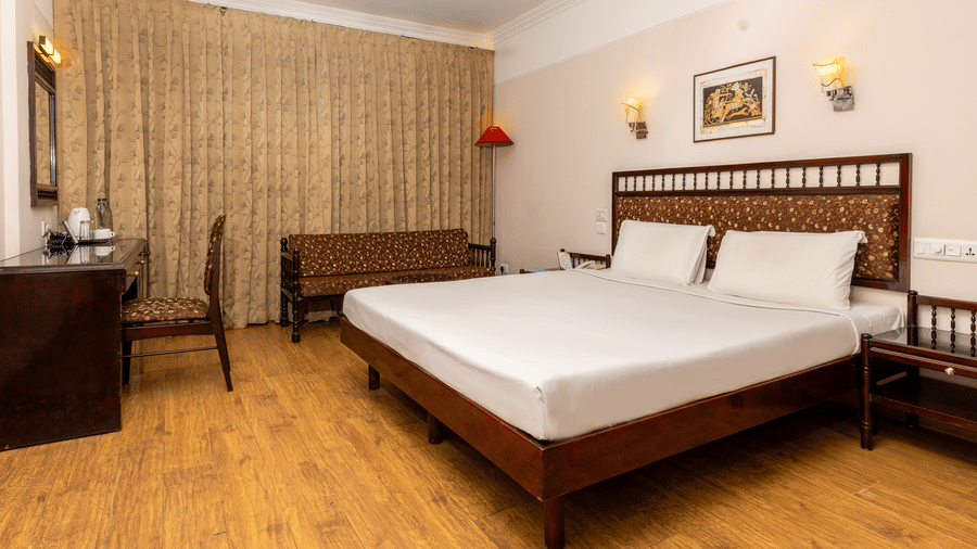 A room with a queen size bed, light wood floors, a work desk, seating area, and floor-to-ceiling beige curtains at Hotel Horizon, Trivandrum.