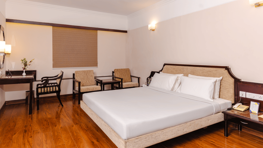 A room with a queen size bed, polished wood floors, 2 armchairs, and a work desk under warm wall lighting at Hotel Horizon, Trivandrum.