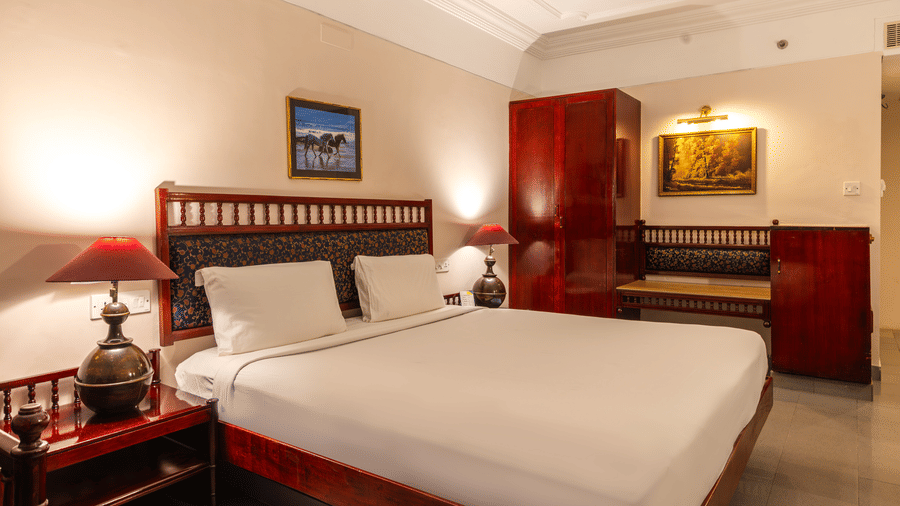A bedroom featuring a queen size bed, dark wood headboard, red bedside lamps, and vintage-style furniture at Hotel Horizon, Trivandrum.