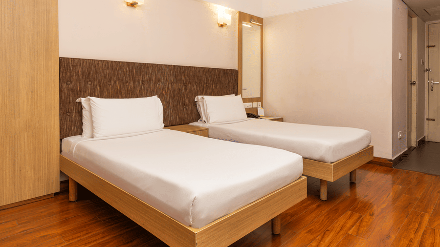 A room featuring twin beds with white linens and brown textured headboards, hardwood flooring, and a wall mirror at Hotel Horizon, Trivandrum.