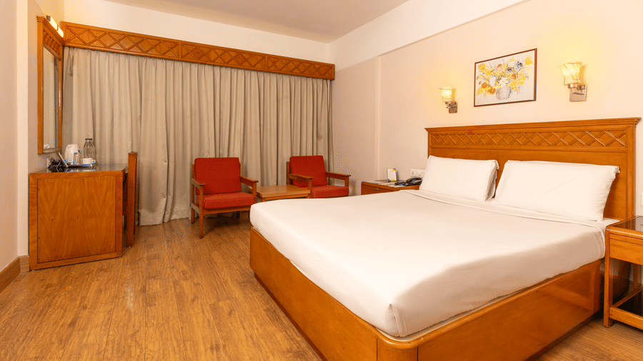 A room with a queen size bed, light wood floors, and a seating area with red armchairs near a window at Hotel Horizon, Trivandrum.