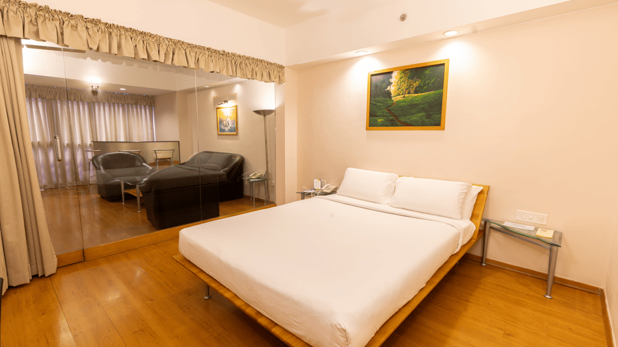 A view of the suite at Hotel Horizon, Trivandrum, featuring a queen size bed, white sheets and pillows, a wall art, and a glass partition showcasing a lounge area beyond.