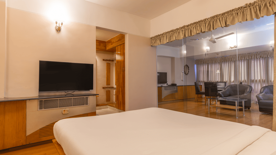 A suite featuring a white bed in the foreground, a wall-mounted TV, and a glass-partitioned lounge behind it at Hotel Horizon, Trivandrum.