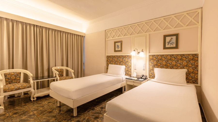 A suite with 2 separate single beds, floral headboards, wall arts, seating area, and white linens at Hotel Horizon, Trivandrum.