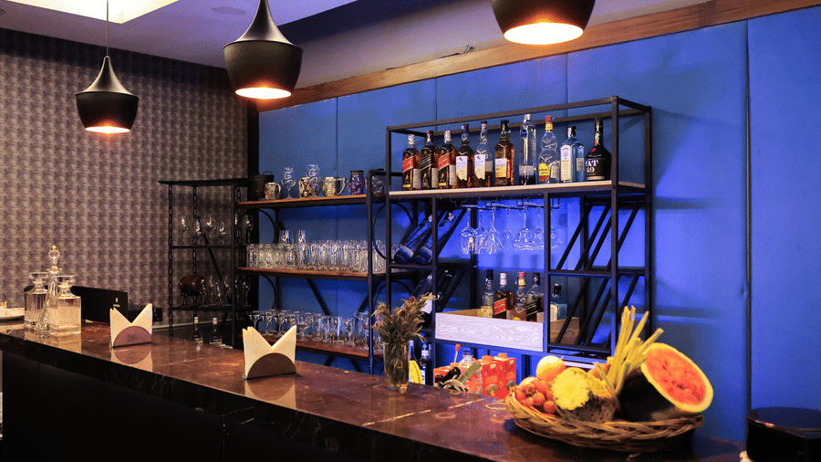 Stylish bar counter with fruit and glasses, a black metal shelving unit, and a blue accent wall with lighting at Hotel Hukam's Lalit Mahal.