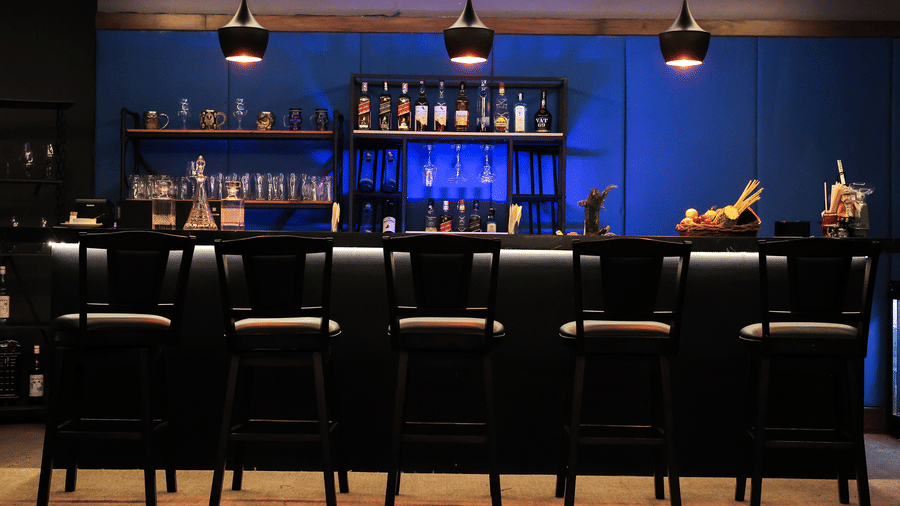 Wide view of the dark, sleek bar with a blue accent wall, shelving full of bottles, and black leather bar stools at Hotel Hukam's Lalit Mahal.