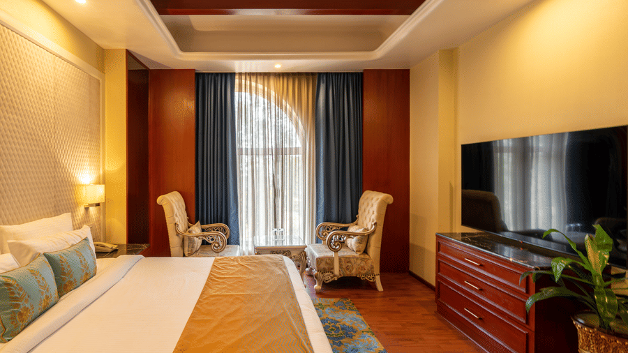 Hotel bedroom featuring a television above a wooden chest, comfortable seating, and a large bed at Hotel Hukam's Lalit Mahal.