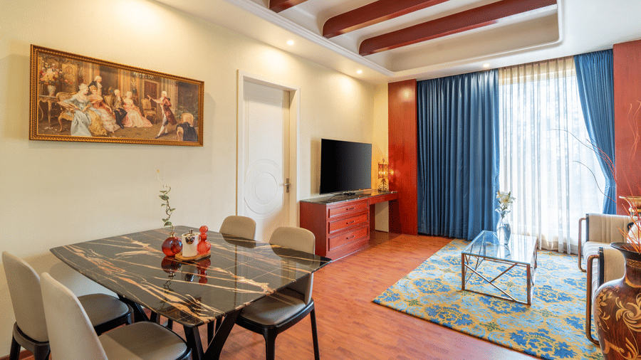 Suite living area with a black dining table, TV, blue rug, and red curtains complementing the wooden ceiling beams at Hotel Hukam's Lalit Mahal.