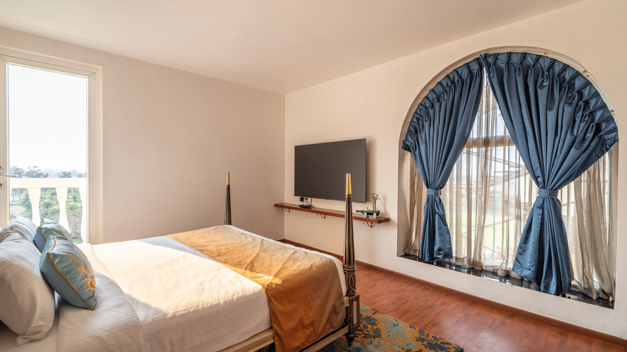 Bright bedroom with a large bed, TV, and blue curtains framing an arch-shaped window overlooking the exterior at Hotel Hukam's Lalit Mahal.
