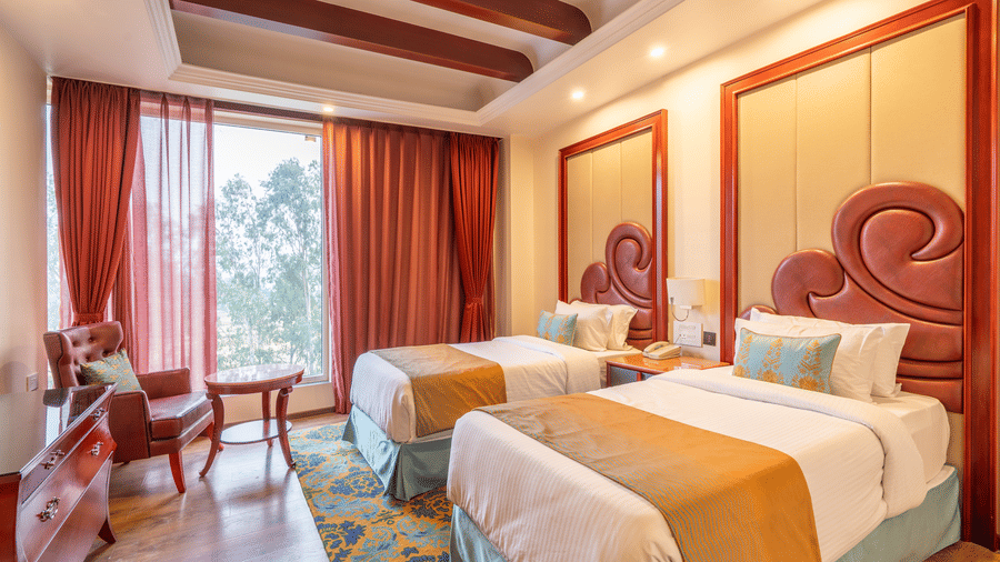 Twin bedroom with rich wooden ceiling beams, gold and blue accents, 2 large beds, and a view through the window at Hotel Hukam's Lalit Mahal.