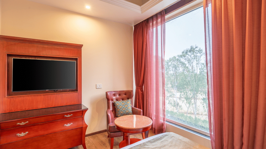 Warmly decorated room featuring a TV, a deep red wooden desk, a chair, and a large window with floor-length curtains at Hotel Hukam's Lalit Mahal.