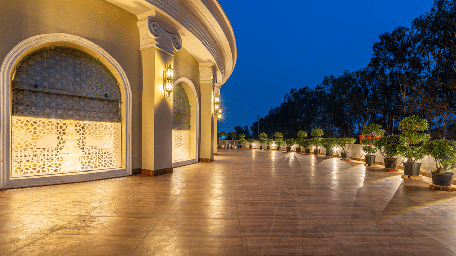 Curved, illuminated walkway at night with decorative wall panels, a polished floor, and potted plants lining the path at Hotel Hukam's Lalit Mahal.